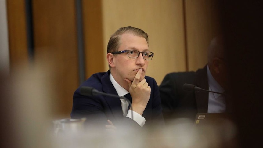 A man in a suit and glasses sits with his hand on his chin at a Senate Committee.