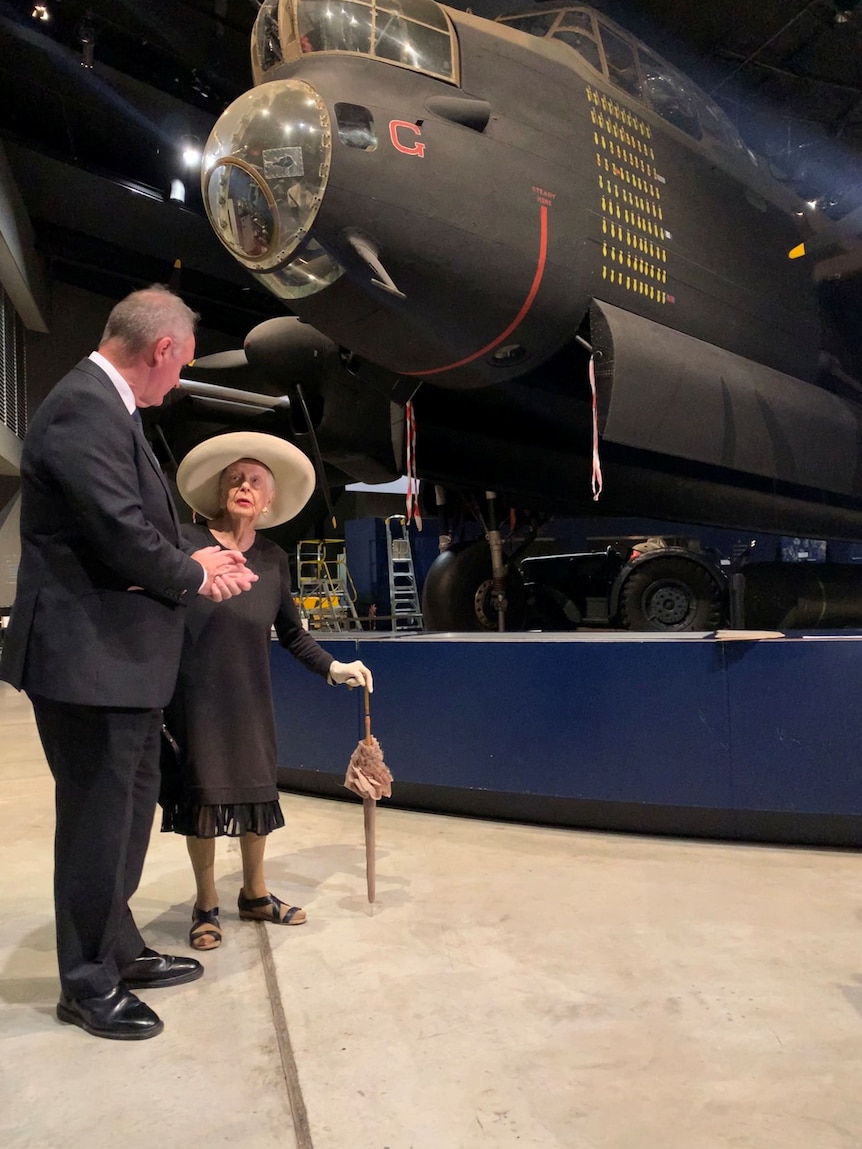 An old woman and a man stand beneath an enormous old fighter plane.