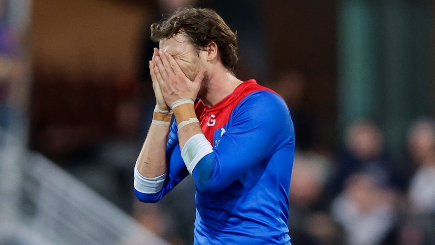 A Demons AFL player holds his face in his hands.