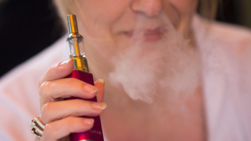 A woman holding an e-cigarette blows smoke through her nostrils.