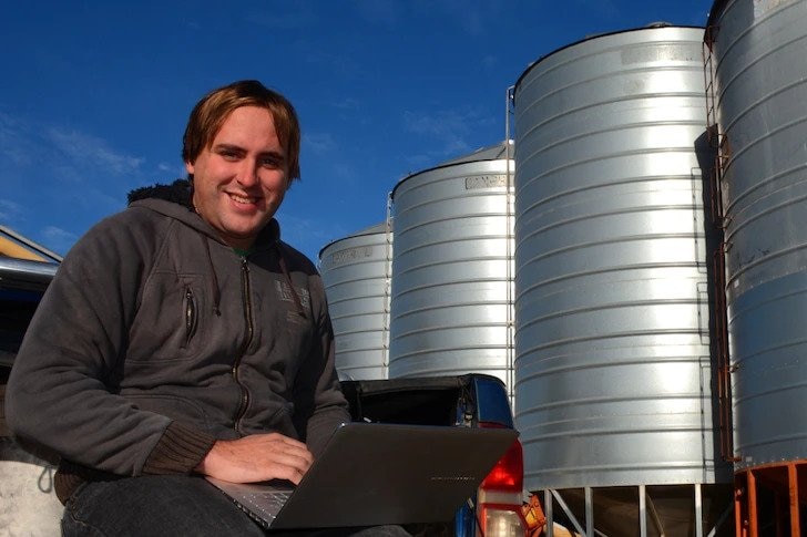 A man sits in the back of his ute looking at a laptop. There are field bins in the background