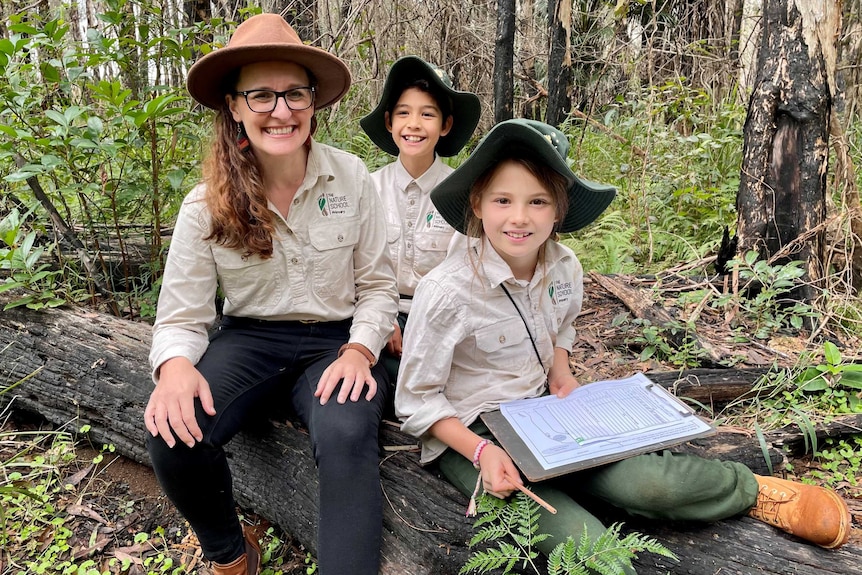 A teacher wearing a broadbrimmed hat sits on a log next to two students.