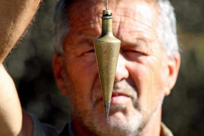 A man with grey hair holds a gold plumb bob in front of his face.