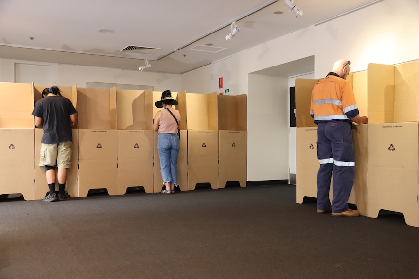 Three people stand with their heads down at cardboard voting booths, as they mark voting ballots, inside a room.