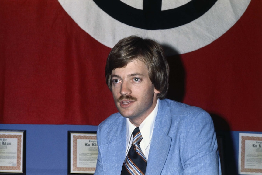 Grand Wizard of the Knights of Ku Klux Klan, David Duke, wears a blue suit and sits in front of a red flag.