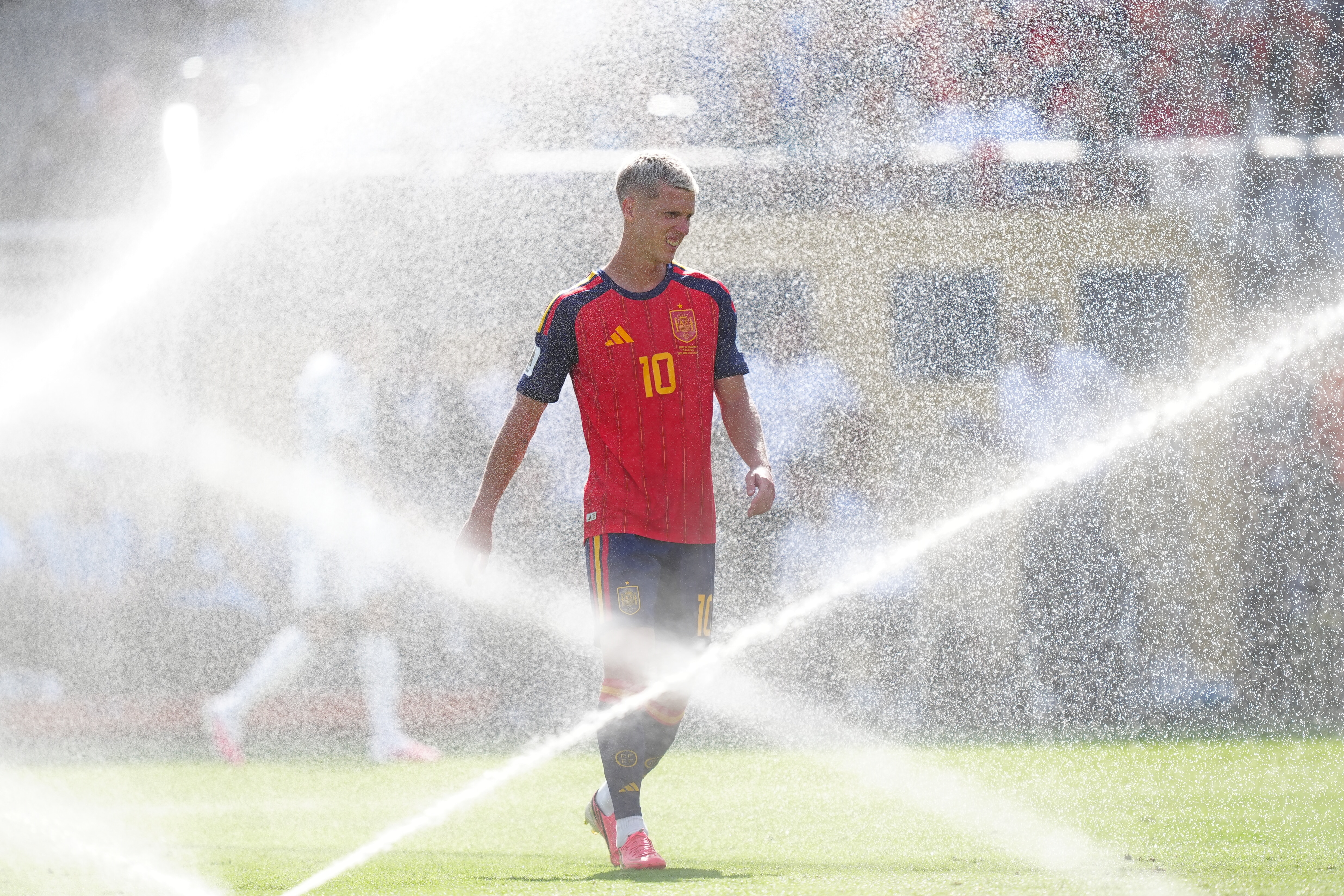 A male soccer player in a red jersey walks through water sprinklers.
