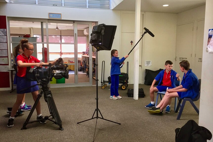 School children operating camera, microphone on boom pole as one student interviews another in classroom.