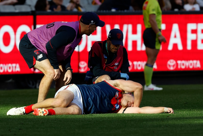 A Melbourne AFL player lies hurt on the ground as two medical staff watch him closely.