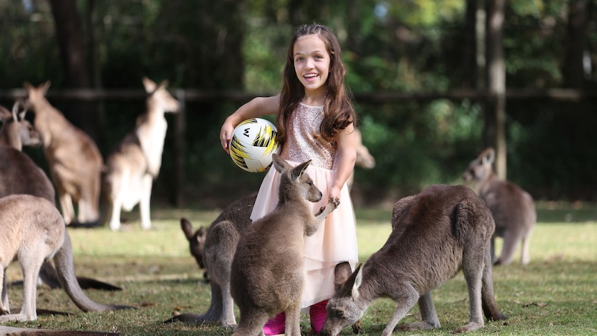a young girl with dwarfism feeds kangaroos at a wildlife reserve. she's smiling at the camera and holding a soccer ball