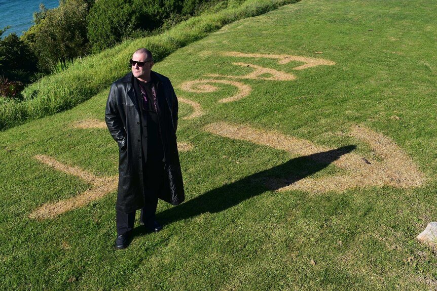 A man in a long black coat and sunglasses standing on green grass that has graffiti burnt into it.