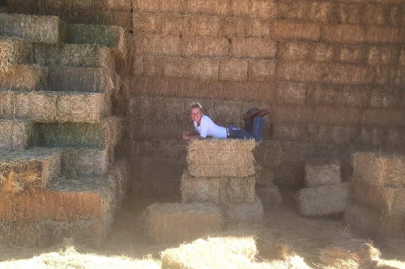 Ruth Hole lying on a hay bale in front of a big stack of bales in their shed at Ulan, NSW.