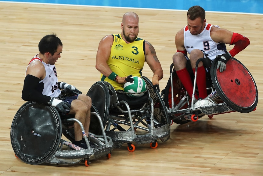 Two US wheelchair rugby stars try to block an Australian with the ball, as one of them comes off one wheel.