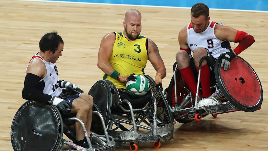 Two US wheelchair rugby stars try to block an Australian with the ball, as one of them comes off one wheel.