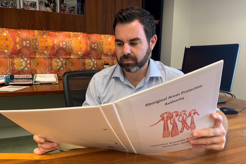 A man sitting at a desk reading a folder labelled Aboriginal Areas Protection Authority