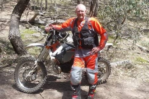 A man wearing orange and black stands next to a trail bike.