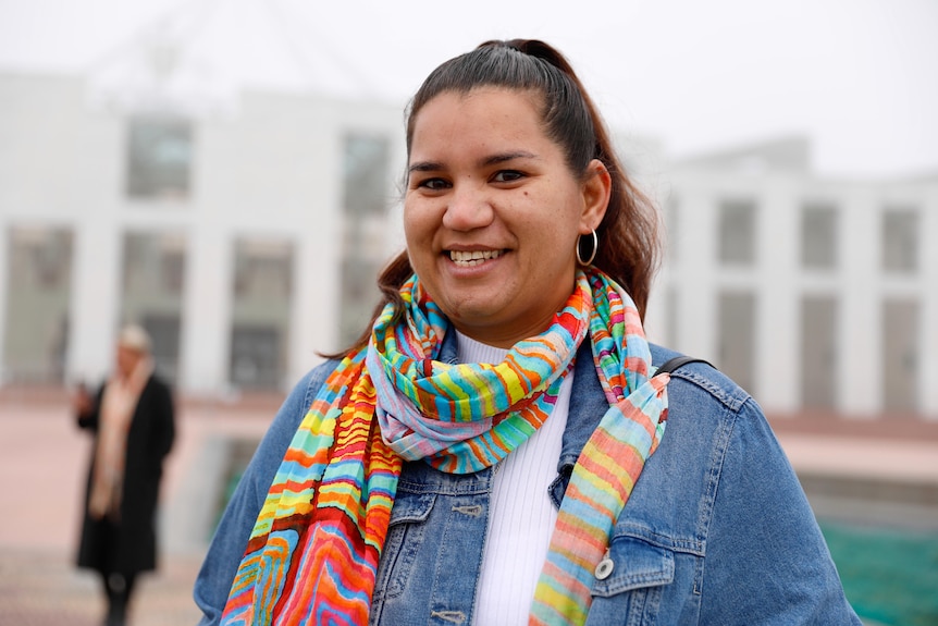 A smiling young Indigenous woman wearing a colourful scarf and denim jacket looks into the camera. 
