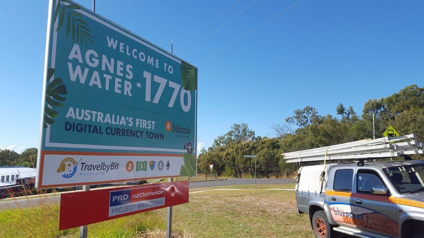 A billboard on a street corner reads 'Welcome to Agnes Water-1770 Australia's first digital currency town'