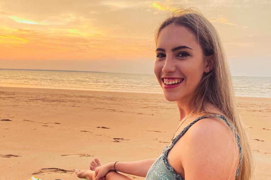 Jennifer Board smiles as she looks back to the camera while sat on the beach at what appears to be sunset.