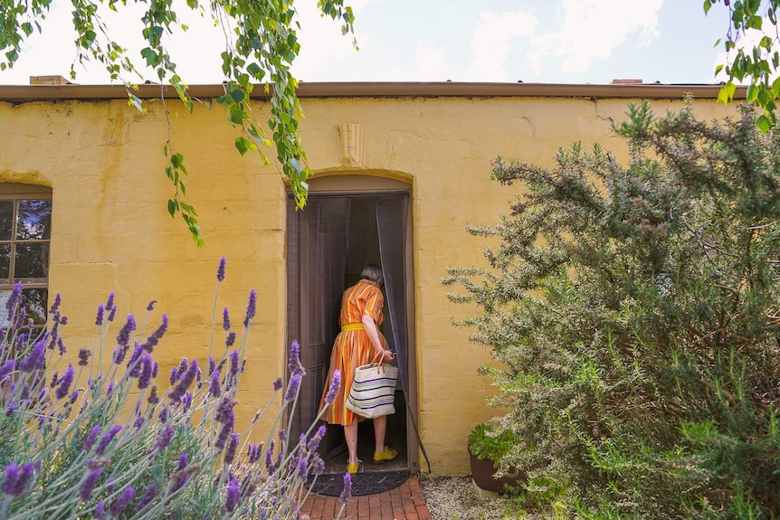 A woman in a brightly coloured frock steps through the door of an old yellow stone heritage building surrounded by lavender bush