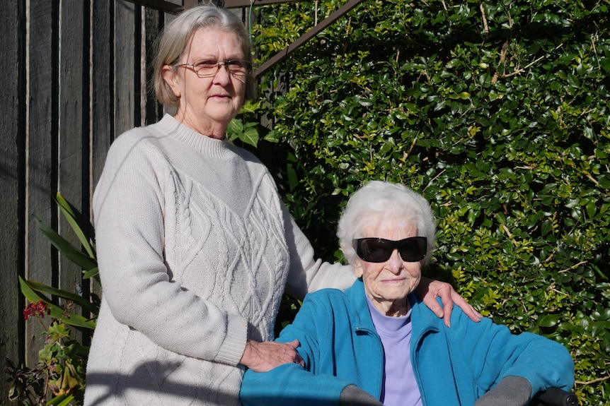 A woman stands next to a sitting elderly woman in a backyard with a green hedge