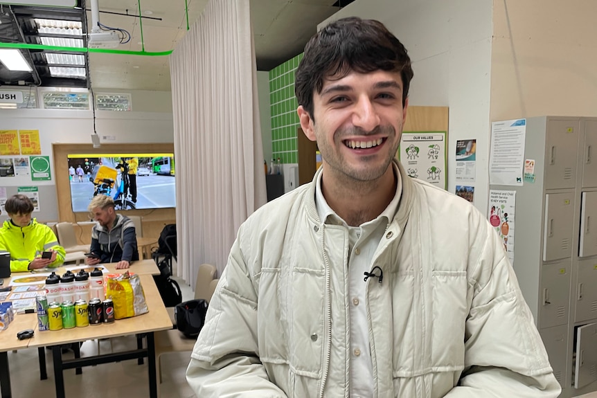 Andrew Copolov smiling at the camera while two delivery riders sit on a table in the background talking