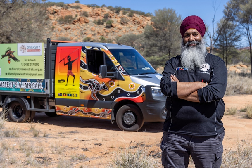 A man in a turban stands in front of a van covered in Indigenous artwork. 