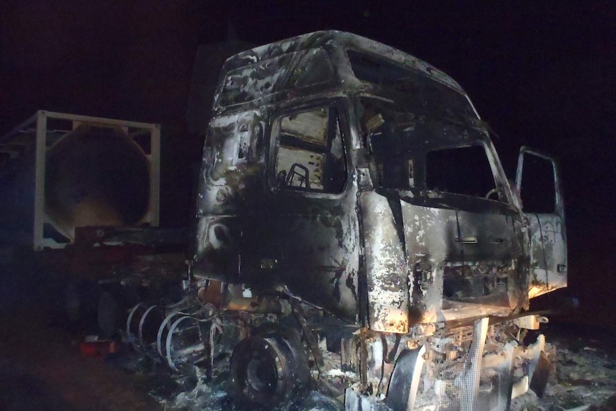 The wreckage of a road train destroyed by fire sits on the side of a highway at night.