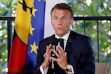 A close up image of the French president with both hands holding a microphone and flags behind him.
