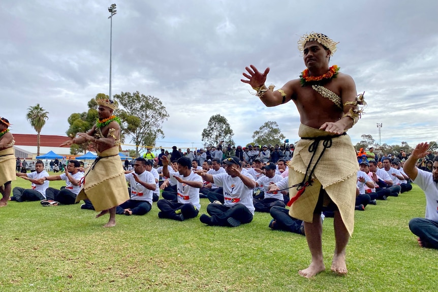 Men in traditional islander dress performing a dance, with rows of cross-legged men behind them