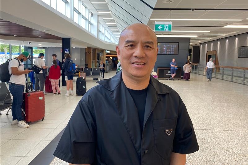 A bald man grins as he poses for a photo inside an aiport arrival area.