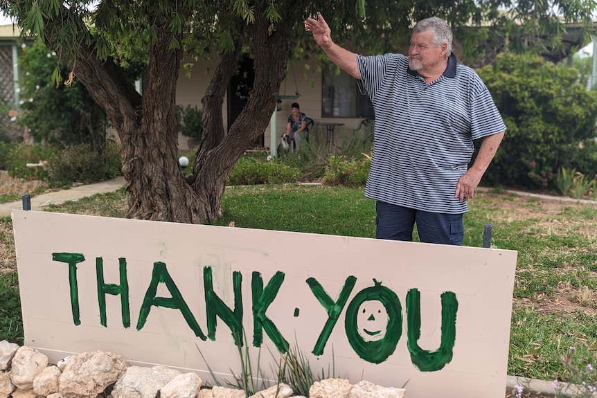 An older white man, John Harding, wearing a navy striped polo stands waving behind a painted sign that says thank you