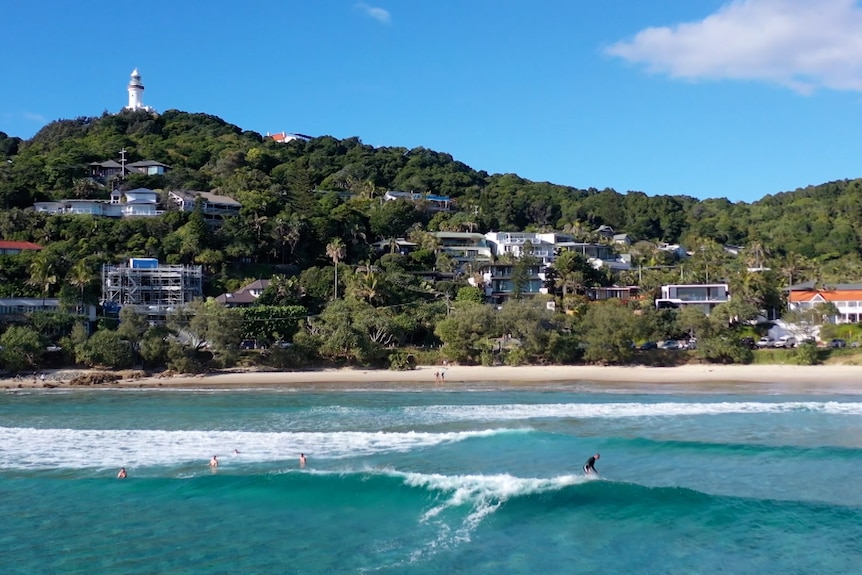 A surfer rides a wave in front of homes beneath a lighthouse