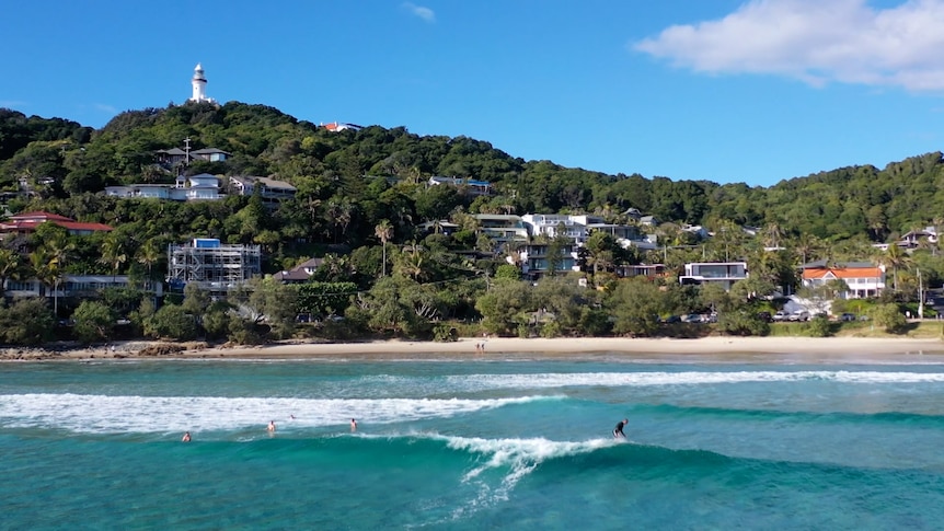 A surfer rides a wave in front of homes beneath a lighthouse