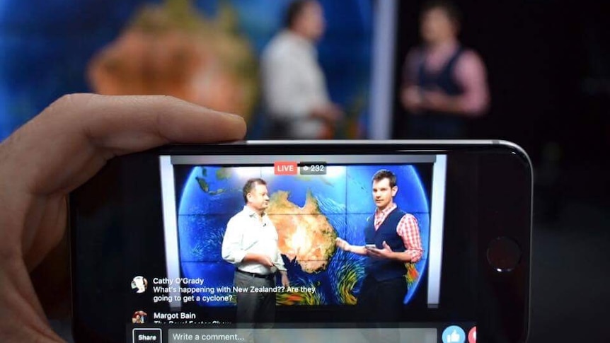 Mobile phone in hand with weather presenters on screen.