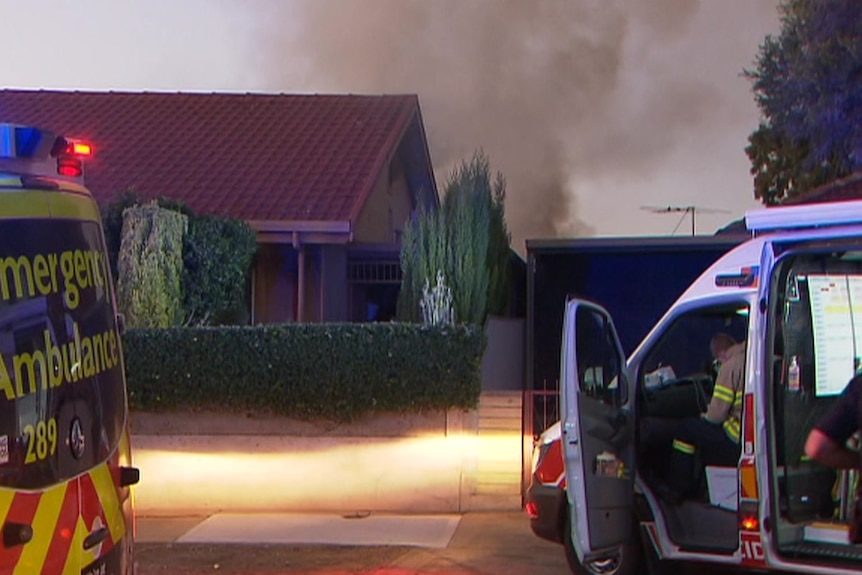 Smoke billows from behind the roof of a suburban home with an ambulance and emergency vehicle parked out front