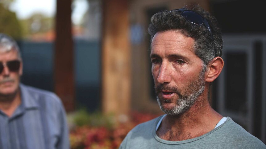 A man with a grey beard and grey t-shirt in front of a building, with a man in sunglasses and a blue shirt in the background.