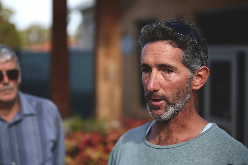 A man with a grey beard and grey t-shirt in front of a building, with a man in sunglasses and a blue shirt in the background.