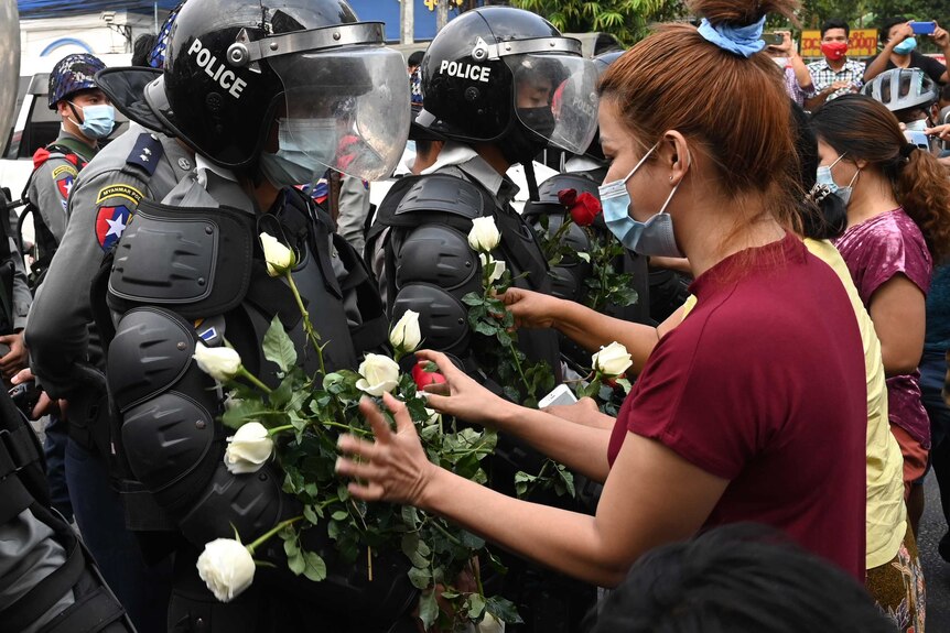 Female protesters wearing face masks place red and white roses onto the armour of riot police in full gear.