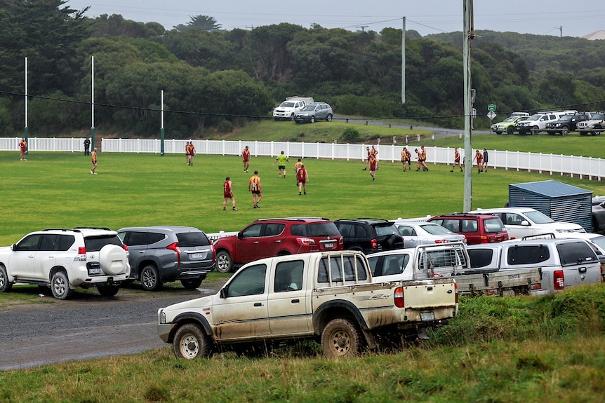 Several cars including a ute on a small hill parked watching over a country football ground with players on field