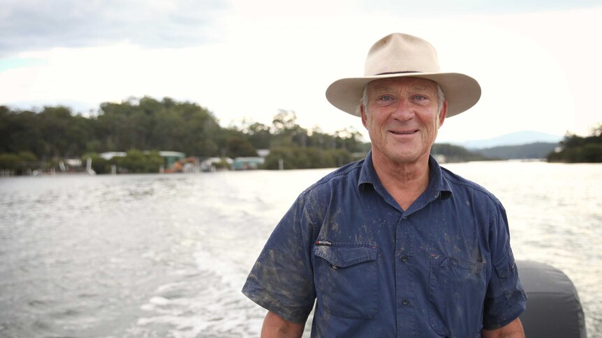 A fisherman wearing an Akubra hat stands on the back of a moving boat.