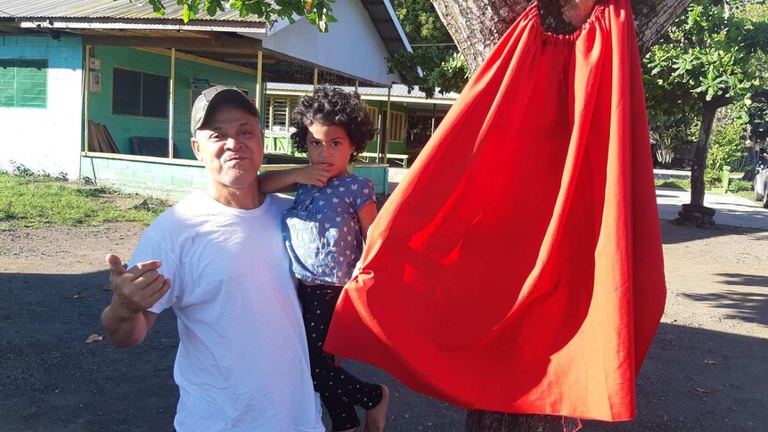 A man holds a young child and stands beside a red flag put up outside a home in Samoa.