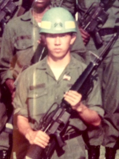 An image of a young man in a green army uniform holding a gun. 