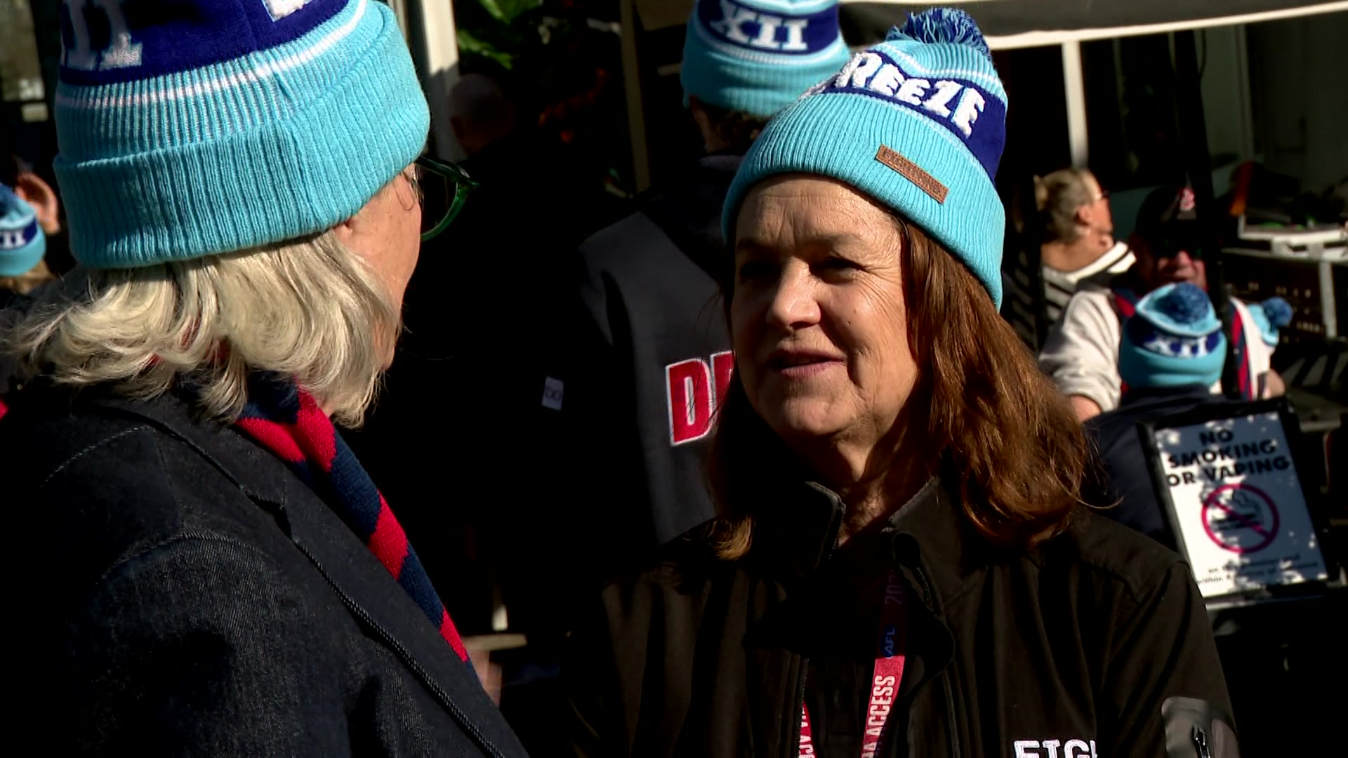 A blonde woman talks to a brunette, both in blue beanies, amongst a crowd.