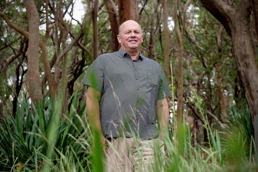 Gary wears a grey button up shirt and shorts with grass in the forecast and forest behind him.