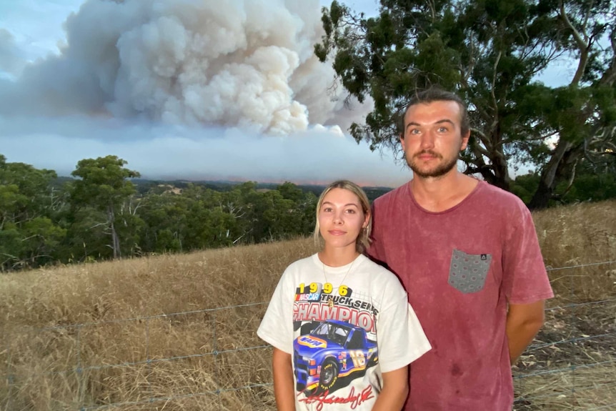 A woman and a man stand in front of trees, a hill and smoke in the sky