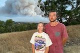 A woman and a man stand in front of trees, a hill and smoke in the sky