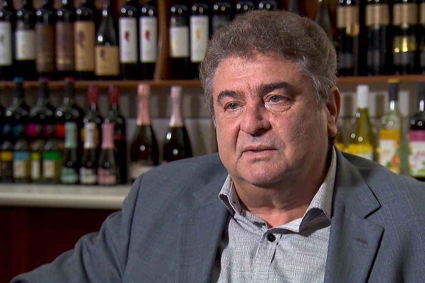 Photo of a man talking in front of wine.
