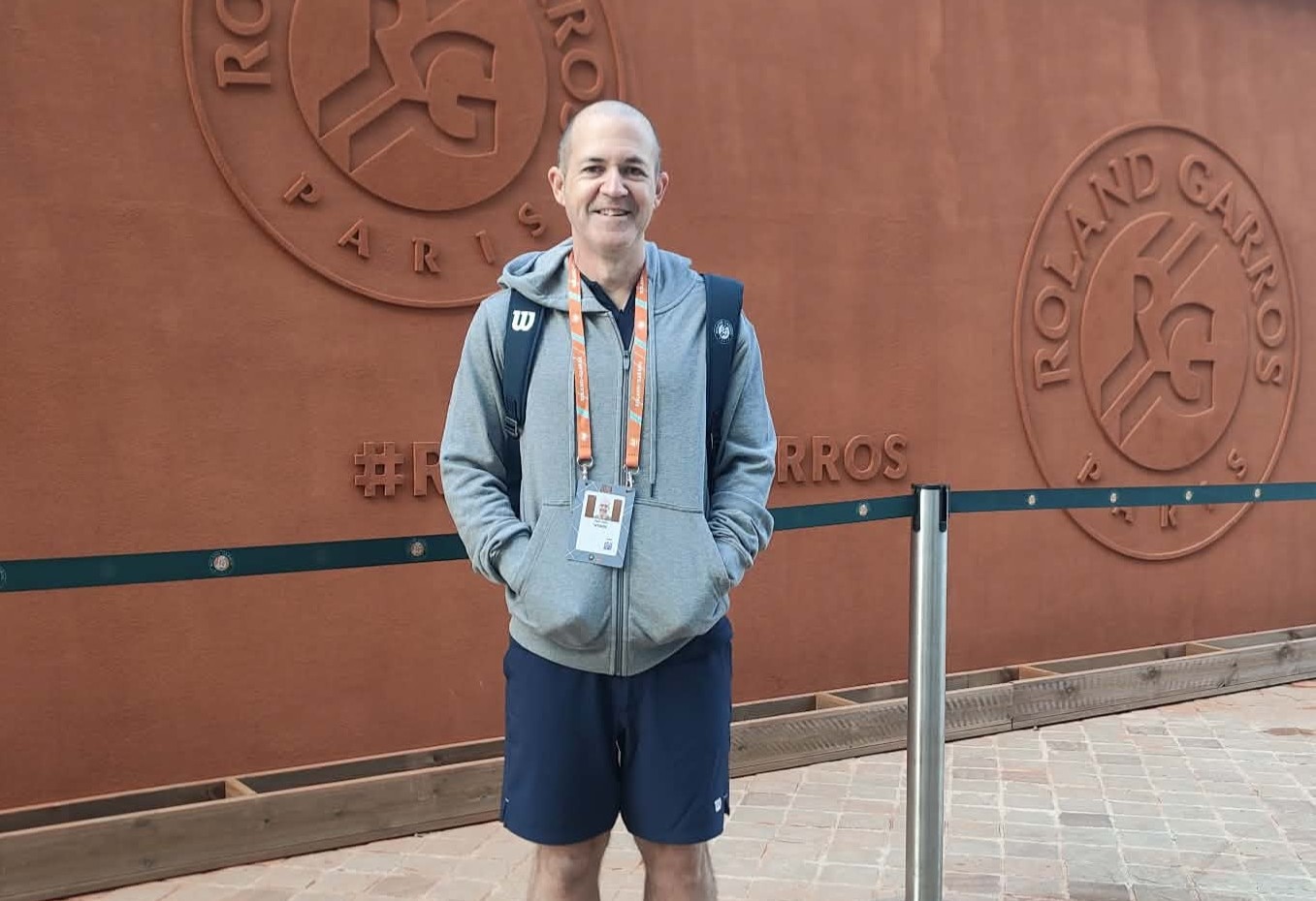 Um homem sorridente vestindo uma camisa fica em frente ao muro de Roland-Garros