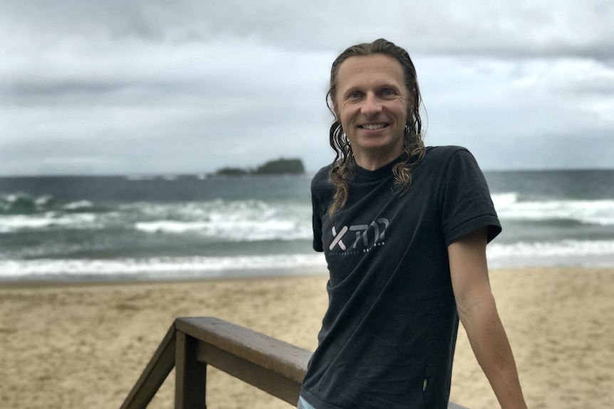 A man leans against a beach boardwalk railing with breaking waves and an island in the background.