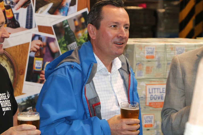 WA Premier Mark McGowan wears a blue jacket and holds a pint of beer, with Gage Roads Brewery imagery in the background.